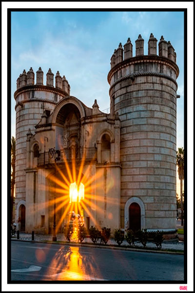 Fotografía al atardecer de la histórica Puerta de Palmas en Badajoz con un espectacular destello de sol dorado brillando a través de su arco central.