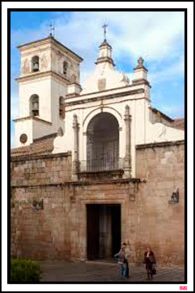 Fachada exterior de piedra de la Concatedral de Santa María la Mayor de Mérida con su torre campanario blanca bajo un cielo despejado.
