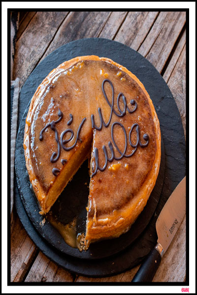 Tarta tradicional Técula Mécula de Olivenza sobre un plato de pizarra, con un corte que muestra su relleno denso de yema y almendra, decorada con letras de chocolate.